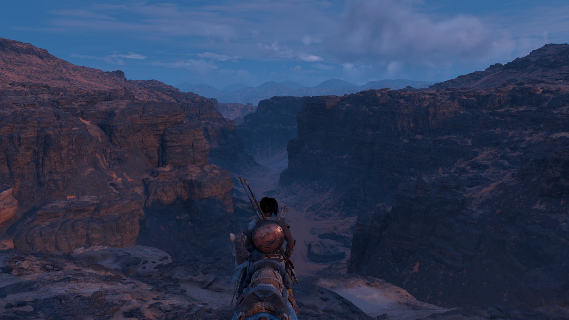 Landscape: a rider sits on a horse on a bluff above an absolutely arid canyon. The top is rocky but the canyon floor is covered in sand.