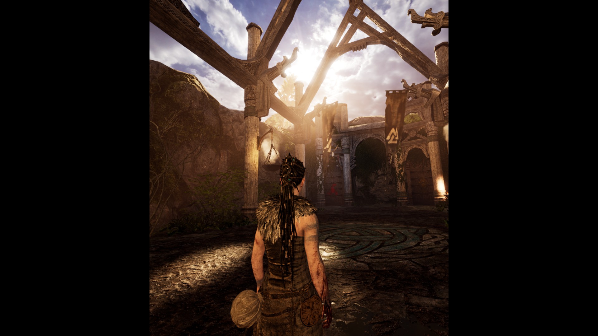 A woman in pictish clothes and tattoos stands in the middle of a broken-down hall. The frame is still standing, and a door at the end, but there's no roof. The sun shines in brightly.